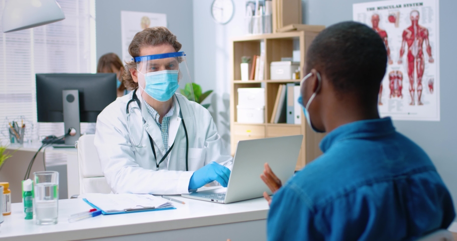Portrait of Caucasian male general practitioner sitting in medical mask and protective face shield in hospital office with African American male patient writing down symptoms and typing on laptop | Shutterstock HD Video #1068813341