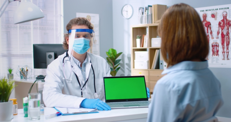 Portrait of male Caucasian physician in protective face shield and medical mask sitting in cabinet talking to woman patient showing laptop with chroma key, monitor with green screen, healthcare | Shutterstock HD Video #1068813359