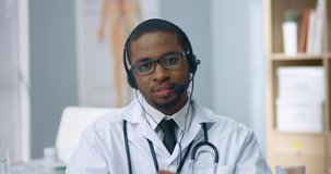 Close up portrait of handsome African American male doctor with stethoscope speaking and explaining in headset with microphone on online consultation sitting in clinic at workplace. Health concept