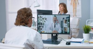 Rear of Caucasian male doctor sitting in hospital speaking on video call with experienced coworker on computer and writing information, training webinar, physician participates in medical conference
