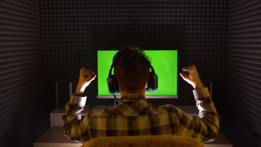 Young adult man in headphones sits before computer or TV screen in poorly illuminated home dark room before chromakey green screen. Concept of rejoicing because of team winning in sports game match.  Royalty-Free Stock Footage #1068821915