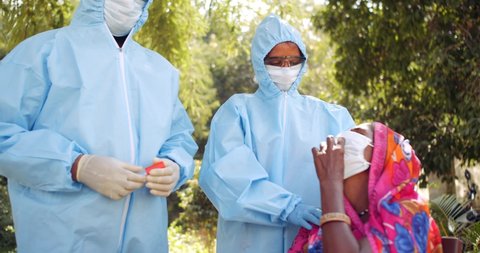 Slow-motion of medical staff in personal protective gear take swap samples from throat mouth of an elderly woman in red saree for virus examination collection with safety workwear in outdoors India