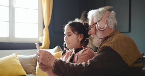 Funny grandfather with his granddaughter uses a mobile phone for a video call with his family. Senior man uses children's pink headphones while playing with a little girl. Family fun at leisure