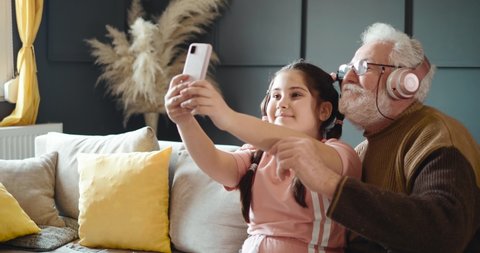 Fun grandfather with headphones taking a picture with his granddaughter. An older man shoots his mouth for a selfie picture with a little girl. Funny grandpa