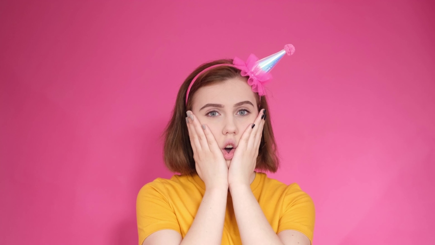 a close-up portrait of a smiling young woman, a girl in a pink holiday cap, clapping her hands with joy on her birthday, experiencing emotions of surprise, amazement.