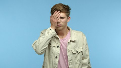 Slow motion of skeptical man facepalm and shake head, staring at camera bothered and annoyed, standing over blue background