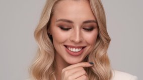 A close-up view of a smiling young blonde woman is looking to the camera while touching her hair standing isolated over gray background in studio - Powered by Shutterstock - Get 15% off with code: PIKWIZARD15