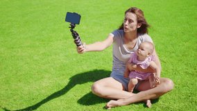 Mother blogger with little daughter having fun records a video blog on green grass.