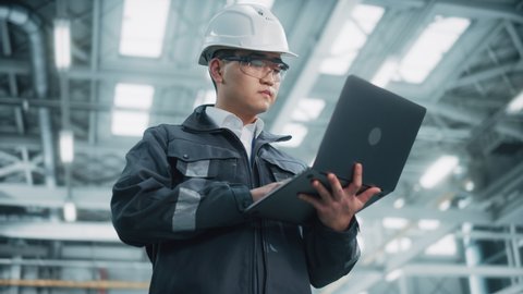 Portrait of a Professional Heavy Industry Asian Engineer Worker Wearing Safety Uniform and Hard Hat Uses Laptop Computer. Confident Chinese Industrial Specialist Standing in a Factory Facility.