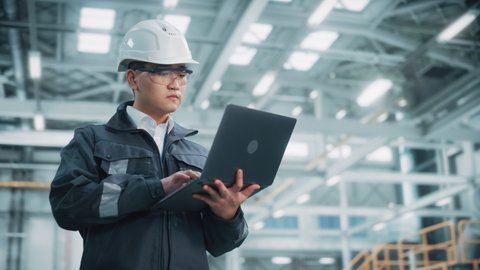 Portrait of a Professional Heavy Industry Asian Engineer Worker Wearing Safety Uniform and Hard Hat Uses Laptop Computer. Confident Chinese Industrial Specialist Standing in a Factory Facility.