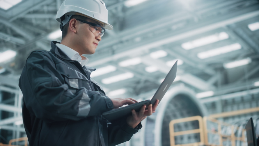 Portrait of a Professional Heavy Industry Asian Engineer Worker Wearing Safety Uniform and Hard Hat Uses Laptop Computer. Confident Chinese Industrial Specialist Standing in a Factory Facility. | Shutterstock HD Video #1068840020