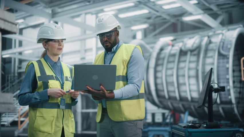 Two Diverse Professional Heavy Industry Engineers Wearing Safety Uniform and Hard Hats Working on Laptop Computer. African American Technician and Female Worker Talking on a Meeting in a Factory. Royalty-Free Stock Footage #1068840050