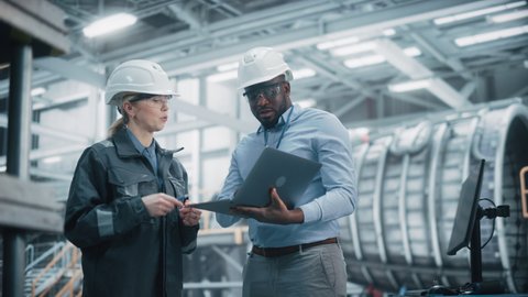 Two Diverse Professional Heavy Industry Engineers Wearing Safety Uniform and Hard Hats Working on Laptop Computer. African American Technician and Female Worker Talking on a Meeting in a Factory.