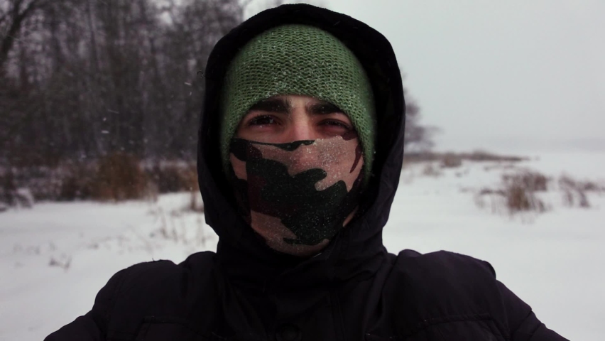 Selfie of young man wearing hood and scarve covering his face who closes his eyes because of snow flying into him. Portrait of a male person standing in wood among falling snowflakes. Theme of tourism
