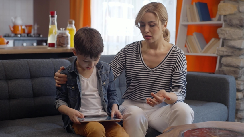 Young woman sitting on the sofa, annoyed by the bad behavior of her little son. She gets angry with her little son, who constantly spends time with a tablet computer. Technology addiction concept.  Royalty-Free Stock Footage #1068843680