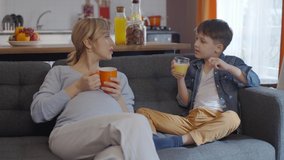 Pregnant woman is chatting while drinking coffee or tea, her little son is drinking juice. Slow motion video.