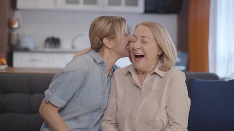 Adult girl sitting on the sofa together in the living room says something secret to her mother's ear. Daughter and mother laugh a lot at what he says. 