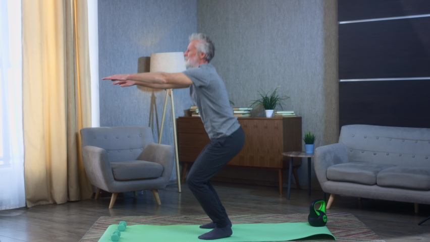 Portrait of a gray-haired senior man squatting on a yoga mat. Home fitness exercise squats on his feet. Grandfather in excellent athletic body shape. Old man in sportswear. Health in old age. | Shutterstock HD Video #1068847187