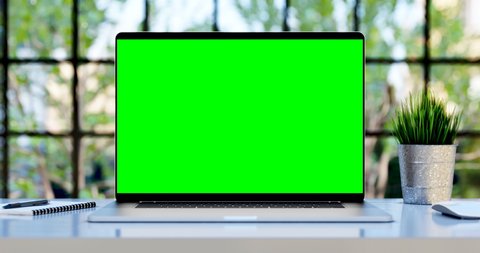 Modern laptop with blank green screen. Static footage with trees swaying or moving in the wind. Home interior or loft office background, 4k 24fps UHD, loop video