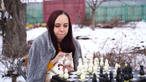 Young woman sitting with cat and playing chess in yard. Female wrapped in grey plaid sitting on street with pet and playing in board game in winter season.