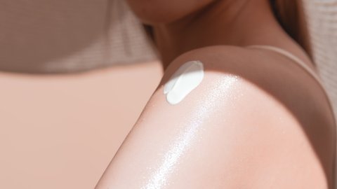 Young slim brown-haired woman in a big white hat thoroughly applies sunscreen on her shoulder against beige background | Sunscreen application shot for body care commercial