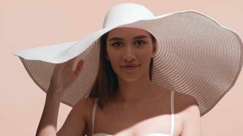 Young good-looking brunette female model in a big white hat covers her face from the sun with her palm looking upward against beige background | Sun cream commercial