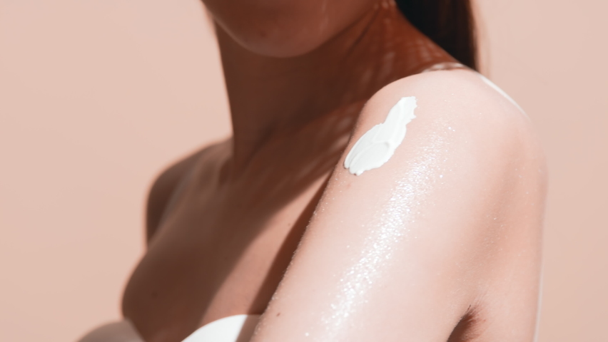 Young good-looking European woman with long dark hair in a straw hat and white bikini applies spf cream on her shoulder smiling and enjoys the sun against beige background | Sunblock commercial | Shutterstock HD Video #1068852230