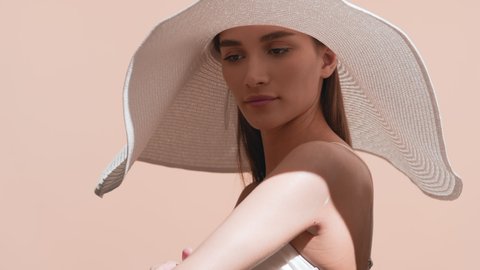 Young pretty European woman with long brown hair in a big white hat and white bikini applies sun cream on her body and enjoys the sun touching her hat against beige background | Sunscreen commercial Video stock