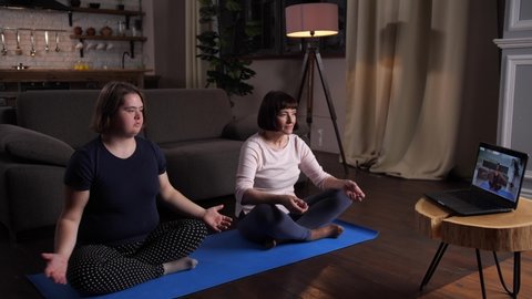 Plus size teen girl with down syndrome and her mother doing yoga exercises using online lessons on laptop. Active woman with disabled daughter practicing lotus position sitting on yoga mat at home