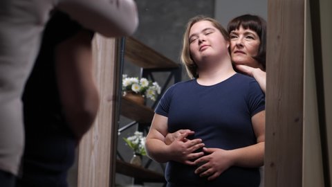 Reflection in mirror of mature woman hugging from back overweight teenage daughter with down syndrome. Caring mother cuddling with curvy handicapped child standing in front of full length mirror