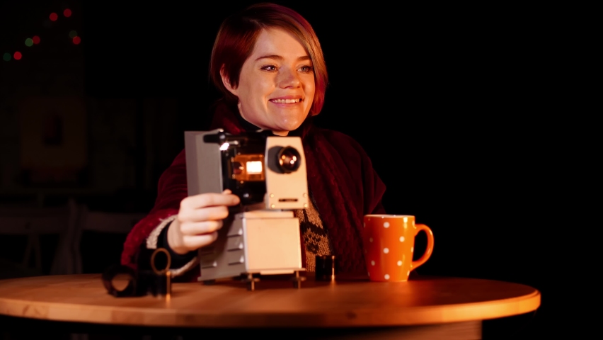 Smiling woman is sitting at the table and watching slides with the help of retro projector. Young caucasian lady spins a wheel to move the film with images. Vintage device for showing photo negatives.