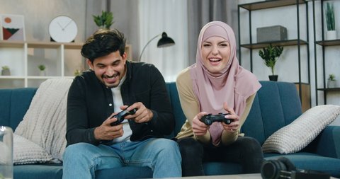 Handsome satisfied bearded guy celebrating victory in video game while his arabic girlfriend in hijab is sad becouse she lost the game,leisure concept