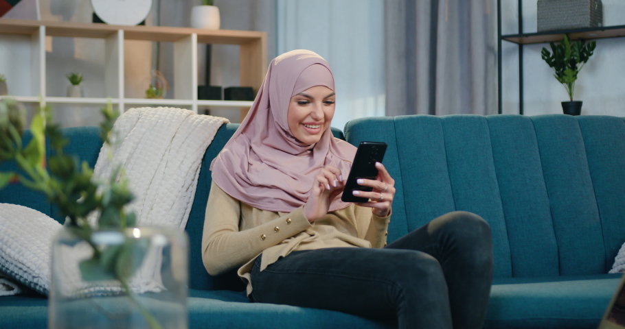 Beautiful in high spirits smiling young woman in hijab and casual clothes sitting on comfortable sofa at home and uses her smartphone,front view | Shutterstock HD Video #1068863717