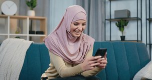 Gorgeous satisfied smiling young arabic woman in hijab sitting on the couch at home and browsing funny photos she got from friends,front view