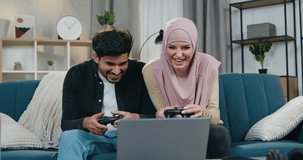 Good-looking satisfied funny multiehnic couple having fun together while playing video games,woman in hijab celebrating victory,bearded guy is upset becouse he lost