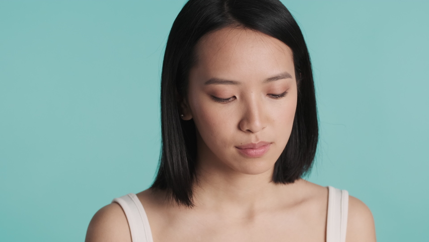 Portrait of young beautiful Asian woman applying cream on face during morning skincare routine over blue background. Beauty concept | Shutterstock HD Video #1068863942