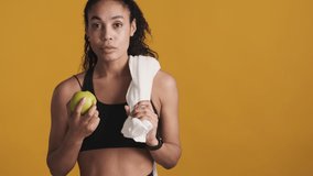 Young attractive Afro woman dressed in sport bra with towel on shoulder eating apple after training over colorful background. Healthy lifestyle concept. Space for text
