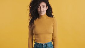Young stylish African American woman with dark fluffy hair dressed in casual wear looking happy waving hello to friend over orange background. Greeting expression