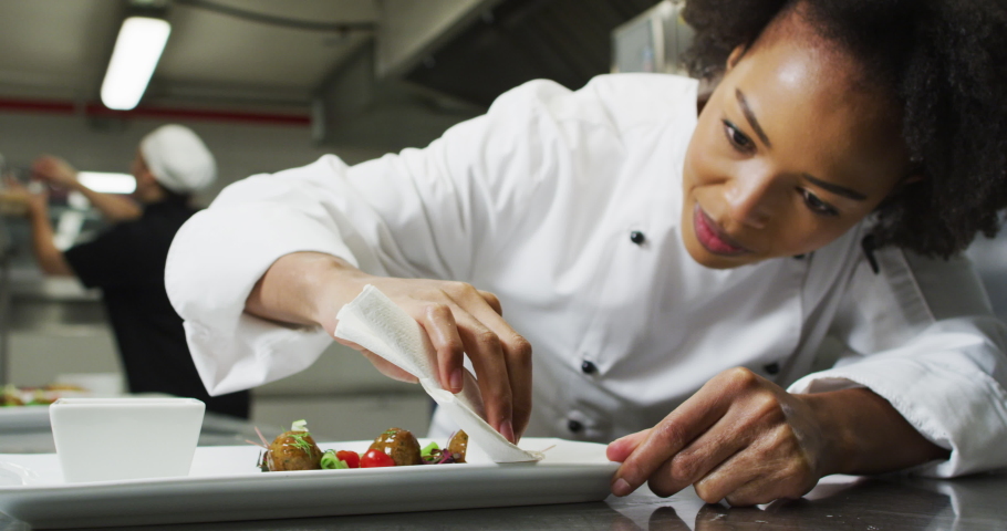 Portrait of african american female chef garnishing dish looking at camera and similing. Working in a busy restaurant kitchen. Royalty-Free Stock Footage #1068866156