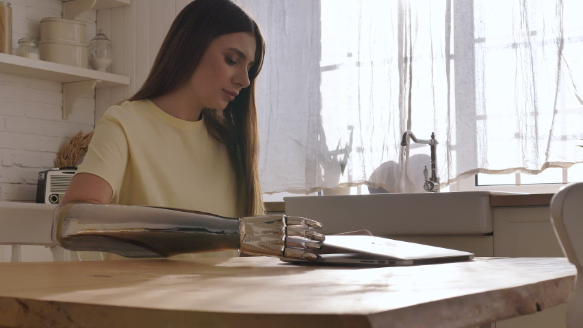 Concentrated woman opens contemporary laptop using shiny bionic arm to surf internet sitting at wooden table in light kitchen