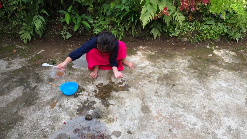 Adorable beautiful cute asian little girl wearing nice dress is enjoy playing water soil stone with plastic bowl at the house yard ground with joy happy, fun during corona covid pandemic new normal.
