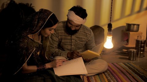 Middle-aged Indian farmer helping his wife to study - adult education. Medium shot of traditional village home with two adults studying together - literacy learning. Indian village