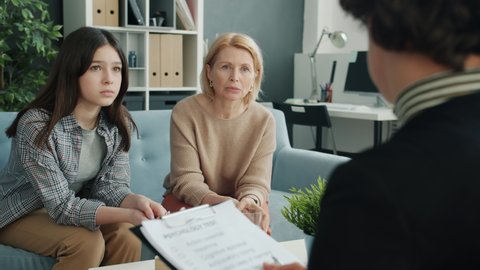 Mature woman is yelling at teenage girl in psychologist's office while therapist is listening and writing offering professional advice to troubled family