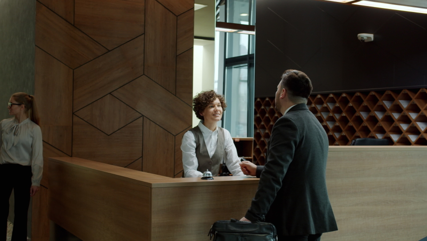 Male guest businessman is talking to hotel receptionist and getting key card while man and woman are walking by. Business accommodation and service concept. Royalty-Free Stock Footage #1068877730