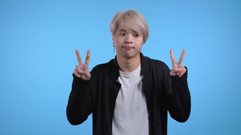 Asian man showing with hands and two fingers air quotes gesture, bend fingers isolated over blue background. Not funny, irony and sarcasm concept.
