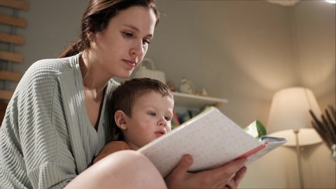 Baby and book, mother and child reading. Woman mom and 2-3 year old boy sit on bed in pajamas in bedroom in evening or night and hold book in hands, looking and reading, turn page. Slow motion