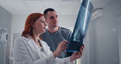 Zoom out view of middle aged woman in medical uniform demonstrating and explaining X ray image to adult man while working in lab in hospital