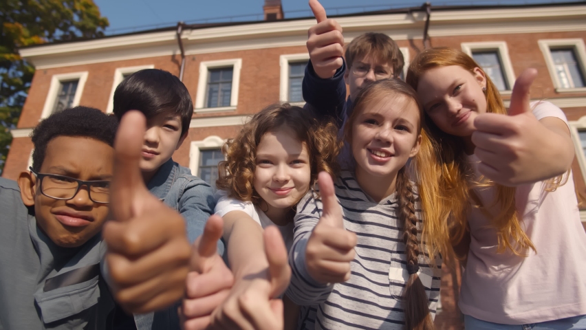 Portrait of happy diverse children looking at camera with thumbs up outdoors. Multiracial school kids gesturing thumb-up smiling at camera outside school building