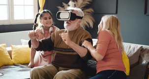 Grandpa uses VR for virtual car driving, grandma and granddaughter laugh. Happy family uses VR glasses