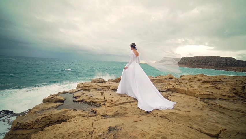Amazing view a young bride in a white wedding dress and veil standing on a rock against the backdrop of sea or ocean and crashing waves Attractive young woman in nature on a background of the landscape.